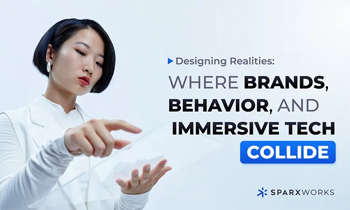 A person with short dark hair and a light complexion, wearing a white collared shirt, appears to be interacting with a translucent digital interface floating between their outstretched hands. The background is a bright, gradient white to light blue. To the right of the person, large, bold text reads "Designing Realities: WHERE BRANDS, BEHAVIOR, AND IMMERSIVE TECH COLLIDE". Below this text is a blue rectangular box containing the word "COLLIDE" in white, bold lettering. At the very bottom right, the Sparxworks logo is visible.
