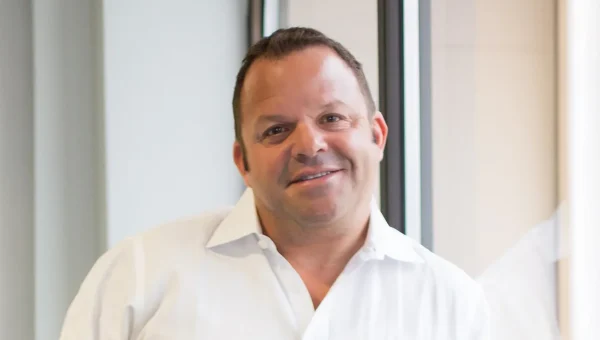 A smiling man in a white dress shirt stands near large windows, facing the camera with a relaxed, approachable business appearance.
