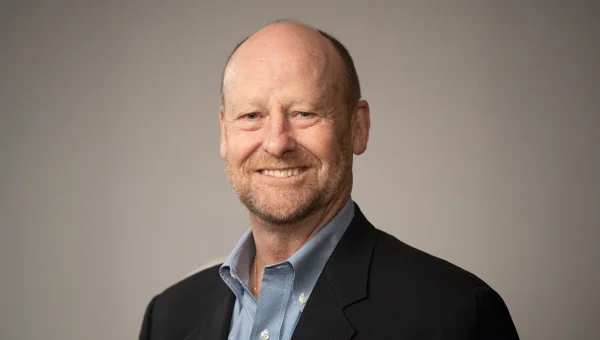 A smiling man with a trimmed beard wears a dark blazer and blue shirt against a neutral background, projecting professionalism.